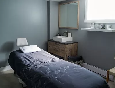 A massage bad in a softy lit room. There is a sin, mirror, window and shelf.