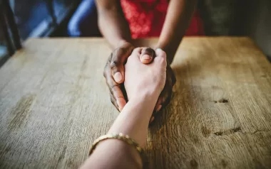 Two people grasping hands in a comforting manner across a table from each other.