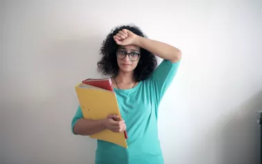 Woman looking distressed holding one arm to her forehead and using the other arm to hold a folder and binder