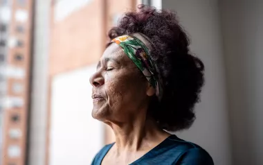 mature woman standing near a window with eyes closed breathing in.