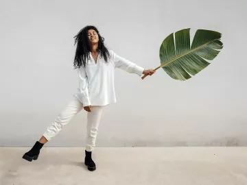 A woman balances on one leg while holding a giant leaf in the other hand, almost as a counterbalance to her raised leg.