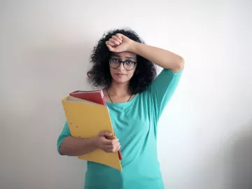 Woman looking distressed holding one arm to her forehead and using the other arm to hold a folder and binder