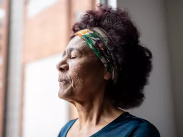 mature woman standing near a window with eyes closed breathing in.