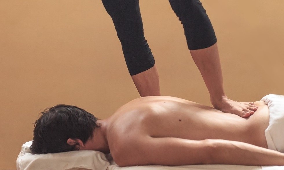 A person lying facedown receiving barefoot massage from a therapist standing over them